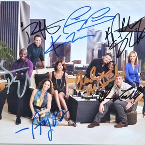May include: A group photo of people posing outdoors, with a cityscape in the background. The photo is signed in multiple colors. The people are dressed in various outfits, and some are seated on a black couch.