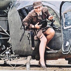 Może przedstawiać: Zdjęcie vintage przedstawiające kobietę w mundurze wojskowym wychodzącą z helikoptera. Ma na sobie brązową kurtkę, spódnicę i kapelusz, z podwiązką i pończochami. Obraz jest podpisany czarnym tuszem.