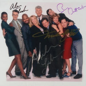 Puede incluir: Una foto grupal de once personas, probablemente un elenco o equipo, posando juntos. La imagen está firmada por varias personas. Las personas visten una variedad de ropa informal de negocios, incluidos trajes, vestidos y blusas. El fondo es blanco liso.