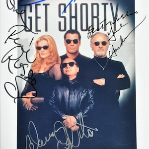Może przedstawiać: Zdjęcie promocyjne filmu "Get Shorty" z czterema aktorami w okularach przeciwsłonecznych. Obraz ma tytuł filmu u góry i wiele podpisów czarnym i niebieskim tuszem. Aktorzy są w ciemnych ubraniach.