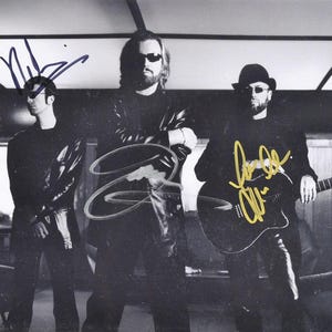 Puede incluir: Fotografía en blanco y negro de un grupo musical. Tres hombres posan, usando gafas de sol y ropa oscura. Uno sostiene una guitarra. La imagen está autografiada con tinta dorada. El fondo es un interior borroso.