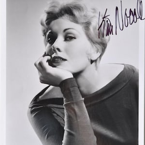 May include: Black and white photograph of a woman with short, wavy hair, resting her chin on her hand. She wears a dark top with long sleeves. The image includes a handwritten signature in the upper right corner.