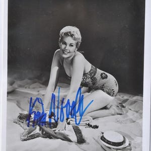 May include: Black and white photograph of a woman in a patterned swimsuit kneeling on a sandy surface. A towel, sunglasses, and a hat are nearby. The image is signed in blue ink.
