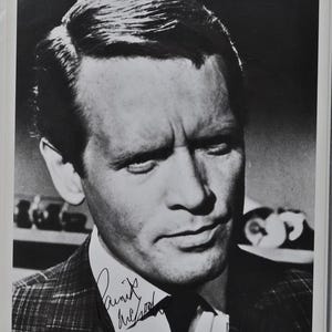 May include: Black and white photograph of a man in a suit, with a plaid pattern. The man has short, neatly styled hair and a serious expression. The photograph includes an autograph in the lower left corner.