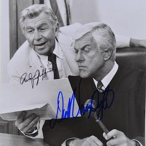 May include: Black and white photograph featuring two men, one in a judge's robe holding a gavel, the other in a suit. Both men are looking at a document. Autographs are visible on the photograph.