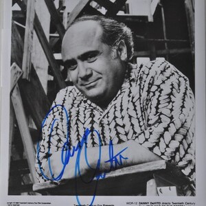 May include: Black and white photograph of Danny DeVito, signed in blue ink. He is wearing a patterned shirt and smiling. The text "THE WAR OF THE ROSES" is visible at the bottom of the image.