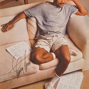 Peut inclure: Une personne assise porte un t-shirt gris à manches courtes, un short kaki, des chaussettes blanches et des baskets blanches. La personne est assise sur un canapé rose clair avec des notes de musique et une signature. La personne se tient la main sur la tête.