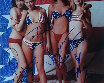 May include: Four women in patriotic-themed swimwear pose in front of an American flag backdrop. The bikinis feature red, white, and blue designs, including stars and stripes. The image is a photograph with signatures on the subjects.