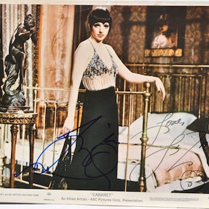 Puede incluir: Foto promocional vintage de la película "Cabaret" con una mujer con top con cuentas y falda negra, apoyada en un marco de cama de latón. La foto incluye firmas y el título de la película.