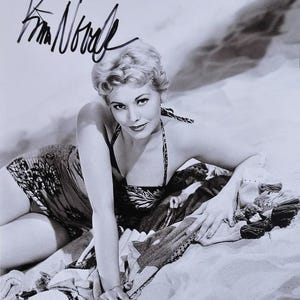 May include: Black and white photograph of Kim Novak, signed in black ink. She is wearing a patterned swimsuit and lying on a beach towel. The image is a vintage portrait.