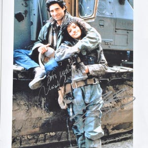 May include: A vintage photograph featuring two people posing together, likely a candid shot. They are dressed in workwear, with one person seated and the other standing, embracing. The image is signed with what appears to be a handwritten autograph.