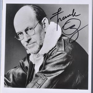 Puede incluir: Fotografía en blanco y negro de un hombre con gafas, camisa blanca y chaqueta de cuero con cuello de borrego. La fotografía está firmada "Frank Oz" con tinta negra.