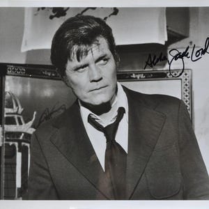 May include: Black and white photograph of a man in a suit and tie, with a white collared shirt. The man is looking off to the side. The photograph is signed "Alana Jack Lord" in black ink.