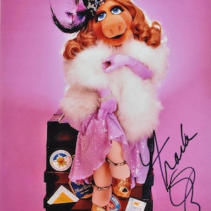 Puede incluir: Miss Piggy, un glamuroso personaje de los Muppets, está sentada en un baúl vintage. Lleva un vestido rosa con lentejuelas, una estola de piel blanca y tacones dorados. Sus accesorios incluyen una pulsera de joyas, guantes y un sombrero con plumas. La imagen está firmada.