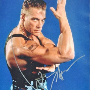 Op de afbeelding: Een kleurenfoto van een man in een zwart tanktop en camouflagebroek, met een Amerikaanse vlag tattoo op zijn arm. Hij buigt zijn arm en er staat een handtekening op de afbeelding.