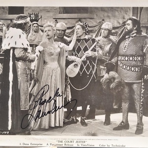 May include: Black and white photograph of a scene from "The Court Jester" featuring actors in period costumes. A woman in a gown is the central figure, surrounded by men in armor and medieval attire. The image includes the text "THE COURT JESTER".