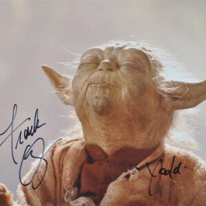 Puede incluir: Primer plano de Yoda de Star Wars, con los ojos cerrados y una ligera sonrisa. Lleva una túnica marrón. La imagen está firmada por Frank Oz y Todd.