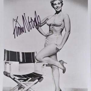 May include: Black and white photograph of a woman in a swimsuit and high heels, posing next to a director's chair. The image is signed with a handwritten signature.