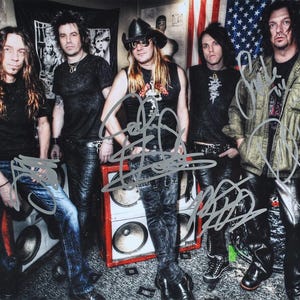 May include: A group of five people, likely a band, posing for a photo. They are dressed in dark clothing, including leather pants and jackets. The background includes an American flag and a speaker cabinet.