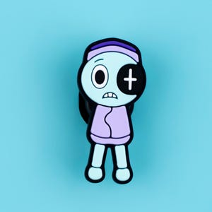 May include: A rubberized shoe charm featuring a cartoon character with light blue skin, a purple headband, and a light purple sweater. The character has one round eye and one black eye patch with a white cross symbol, and a worried facial expression.
