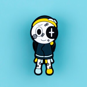 May include: A shoe charm featuring a cartoon character with a white face, one circular eye, and one eye with a cross symbol. The character wears a yellow headband, a dark blue jacket, and has one light blue leg and one yellow leg with grey spots.