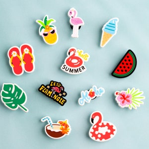 May include: A set of 11 colorful shoe charms featuring summer themes, including a pineapple, flip flops, a watermelon slice, a flamingo, a coconut drink, a flower, and an ice cream cone. The charms have various phrases printed on them, such as "Let's Flamingle", "Aloha", and "Hello Summer".