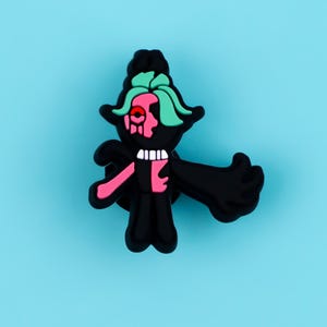 May include: A black shoe charm featuring a stylized character with mint green hair, a pink face section, and a single red eye. The figure has a black body, a white collar, and one extended arm. The design is flat with raised edges and a matte finish.