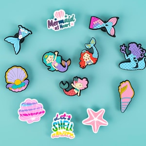 May include: A set of ten colorful shoe charms featuring mermaid and seashell designs. The charms include a mermaid with a crown, a mermaid with a tail, a seashell, a starfish, and a charm that says "Let's Shell-ebrate".
