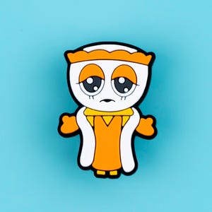 May include: A rubberized pin featuring a cartoon character with a white face, large dark eyes, and orange eyelids. The character wears an orange crown, an orange dress with a yellow collar, and white trim, all outlined in thick black borders.
