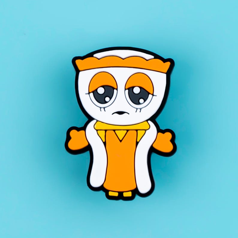May include: A rubberized pin featuring a cartoon character with a white face, large dark eyes, and orange eyelids. The character wears an orange crown, an orange dress with a yellow collar, and white trim, all outlined in thick black borders.