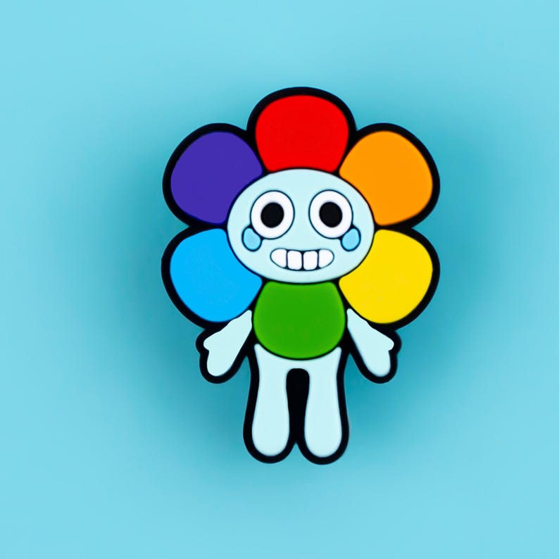 May include: A cartoon character charm featuring a light blue head and body with a wide, toothy grin and large black eyes. The character is framed by a vibrant, multi-colored flower petal border in red, orange, yellow, green, blue, and purple.