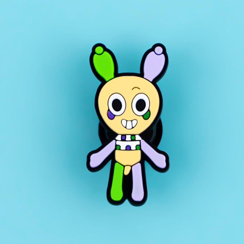 May include: A cartoon character enamel pin featuring a pale yellow head and body with large white eyes, a wide smile, and two tall ears. The character has light green and lavender accents on the ears, cheeks, and limbs, with a black outline.