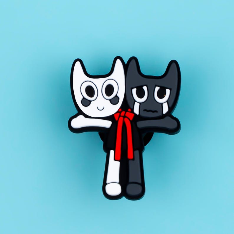May include: A shoe charm featuring two conjoined cartoon figures, one white with a smiling expression and one black with a crying expression, both linked by a bright red ribbon tied around their waists against a solid light blue background.