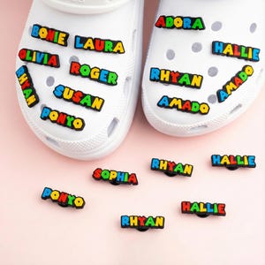 Op de afbeelding: Witte Crocs-schoenen versierd met kleurrijke naam bedels. De bedels spellen namen als "Bonie", "Laura", "Olivia", "Roger", "Susan", "Ponyo", "Adora", "Hallie", "Rhyan" en "Amado". Extra bedels zijn verspreid.