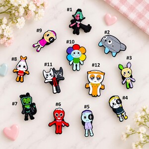 May include: A collection of twelve colorful cartoon-style shoe charms featuring various characters, including a rainbow flower, a grey creature, a red figure, and several humanoid designs, each labeled with a number from one to twelve.