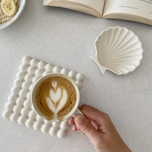 Può includere: Un sottobicchiere quadrato bianco con una texture a bolle in rilievo, un piattino in ceramica bianca a forma di conchiglia e una tazza bianca riempita di caffè con latte art a forma di cuore. Gli oggetti sono vicini a un libro e una ciotola.