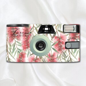 May include: A white disposable camera with a floral design featuring pink flowers and green leaves. The camera has a black lens and a flash. The text "Forever Memories" is printed on the front of the camera.