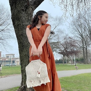 May include: A woman wearing a rust-colored maxi dress holds a white macrame bag with a wooden handle and a tassel. The bag has a woven pattern and a fringe.