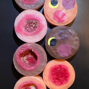 Puede incluir: Seis barras de jabón redondas, de colores vivos y con diseños variados, hechas a mano. Algunas tienen un patrón de espiral, otras tienen un agujero en el centro y otras tienen un patrón de lunares. Los jabones son de diferentes colores, incluyendo rosa, marrón y naranja.