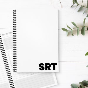 May include: White spiral-bound notebook with the letters "SRT" in bold black font. The notebook is open to reveal lined pages. The words "TODAY WILL BE A GREAT DAY" are visible on a separate page. Green leaves are in the background.