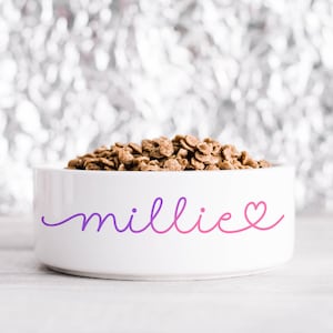 May include: A white ceramic pet bowl filled with brown pet food. The bowl features the name "millie" in a purple to pink gradient script with a heart. The bowl is on a white surface with a blurred silver background.