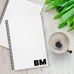 May include: A white spiral-bound notebook with the initials "BM" in bold black letters. A cup of coffee with a gold handle sits to the right, and green leaves are in the background. The notebook is on a light wood-grain surface.