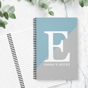 May include: A notebook with a spiral binding, featuring a blue and grey diagonal design. The cover displays a large white letter "E" and the text "EMMA'S NOTES". An open notebook with lined pages is also visible.