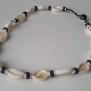 Może przedstawiać: Naszyjnik wykonany z kremowych muszli i czarnych przekładek. Muszle mają owalny kształt i są nawleczone na czarny sznurek. Naszyjnik leży na białej powierzchni.