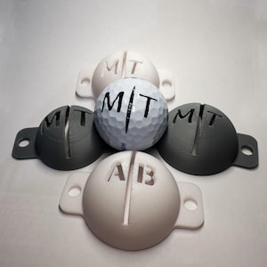 Könnte beinhalten: Vier 3D-gedruckte Golfballhalter, zwei weiße und zwei graue, mit den Buchstaben "MT" auf den grauen Haltern und "AB" auf dem weißen Halter. Ein weißer Golfball mit den Buchstaben "MT" darauf befindet sich in der Mitte der Halter.