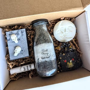 Puede incluir: Una caja de regalo llena de artículos de cuidado personal. Incluye un tarro de sales de baño de naranja dulce, un jabón con forma de calavera, una bomba de baño con forma de luna, un bálsamo labial y una pequeña bolsa negra con una pegatina de agradecimiento.