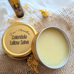 Puede incluir: Un tarro de pomada de caléndula y sebo con tapa dorada, junto a un tarro abierto de la pomada. La tapa tiene el texto "Pine + Clementine Calendula Tallow Salve". También se ve una botella de vidrio marrón y pétalos de flores amarillas secas.