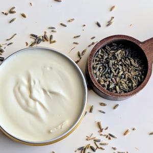 May include: A close-up of a white cream in a round metal tin, alongside a wooden scoop filled with dried lavender buds. Dried lavender buds are scattered on the white surface, suggesting a natural skincare product.