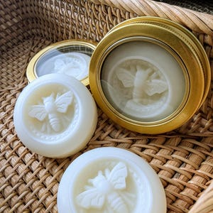 May include: Close-up of several white, round soaps with a bee design, some in gold-rimmed tins. The soaps are arranged in a woven basket, creating a natural and rustic aesthetic. The bee design is a prominent feature.