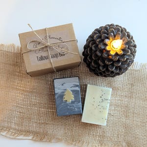 May include: A rectangular cardboard box tied with twine, labeled Pine & Clementine Blue Ridge Tallow Soap Bar, next to a dark brown pinecone with a glowing light and two rectangular soap bars, one grey with a small tree icon and one cream with specks.